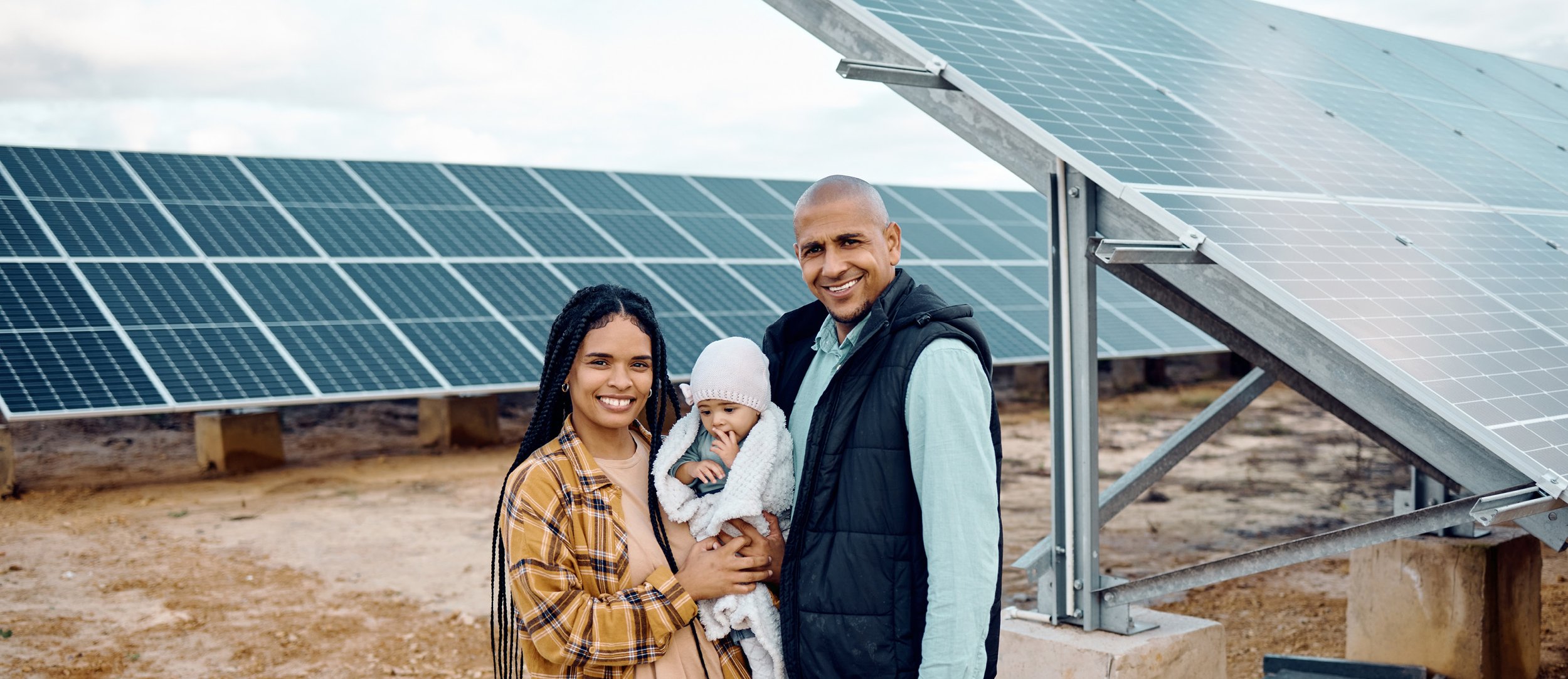Family of three, a woman, a man, and a baby, standing outdoors near solar panels, smiling at the camera.