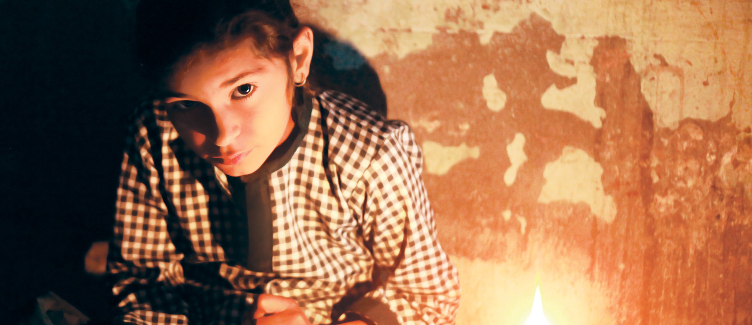 A young girl with light brown hair and blue eyes, wearing a checkered shirt, sitting on the floor beside a lit candle against a textured wall.