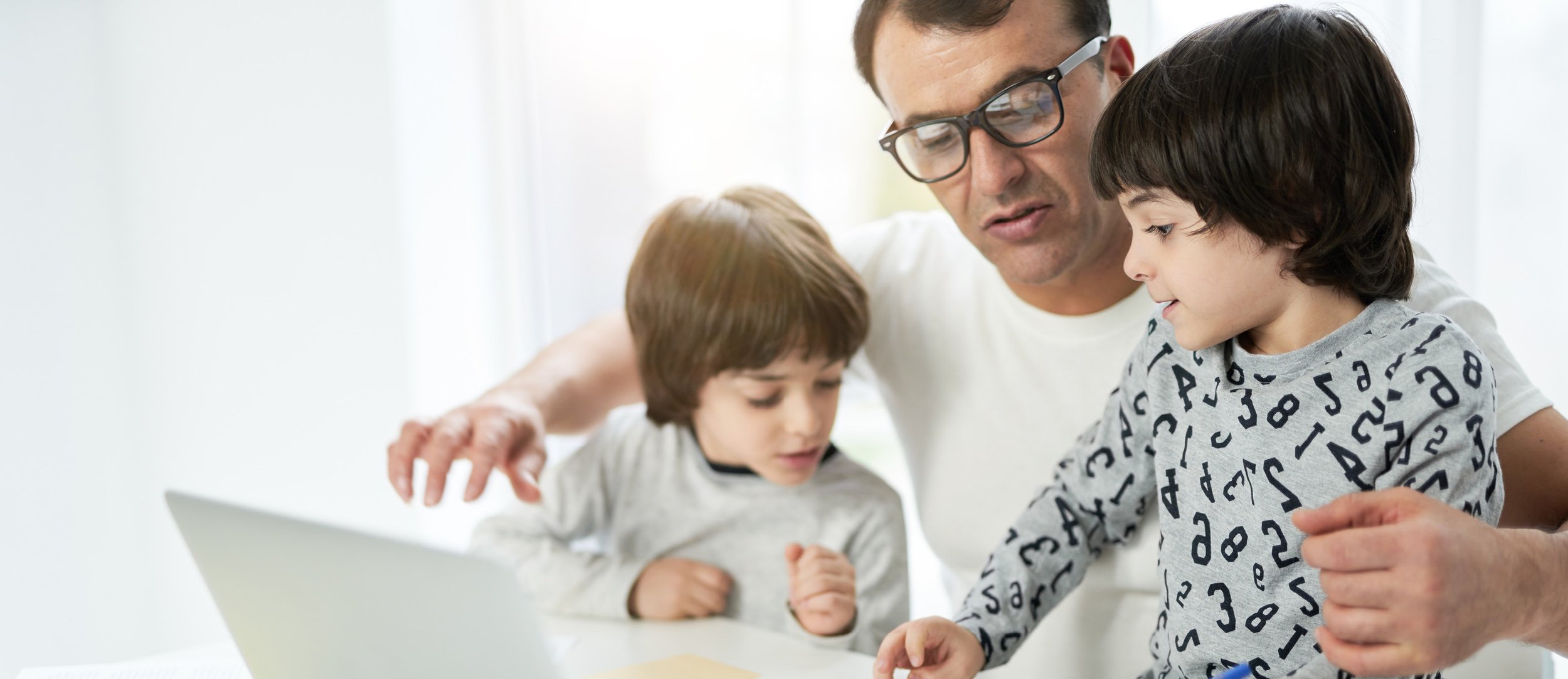 A man wearing glasses and a white t-shirt helping two young boys with a project or activity at a table in a bright room. The boys are focused, and there is a laptop on the table.