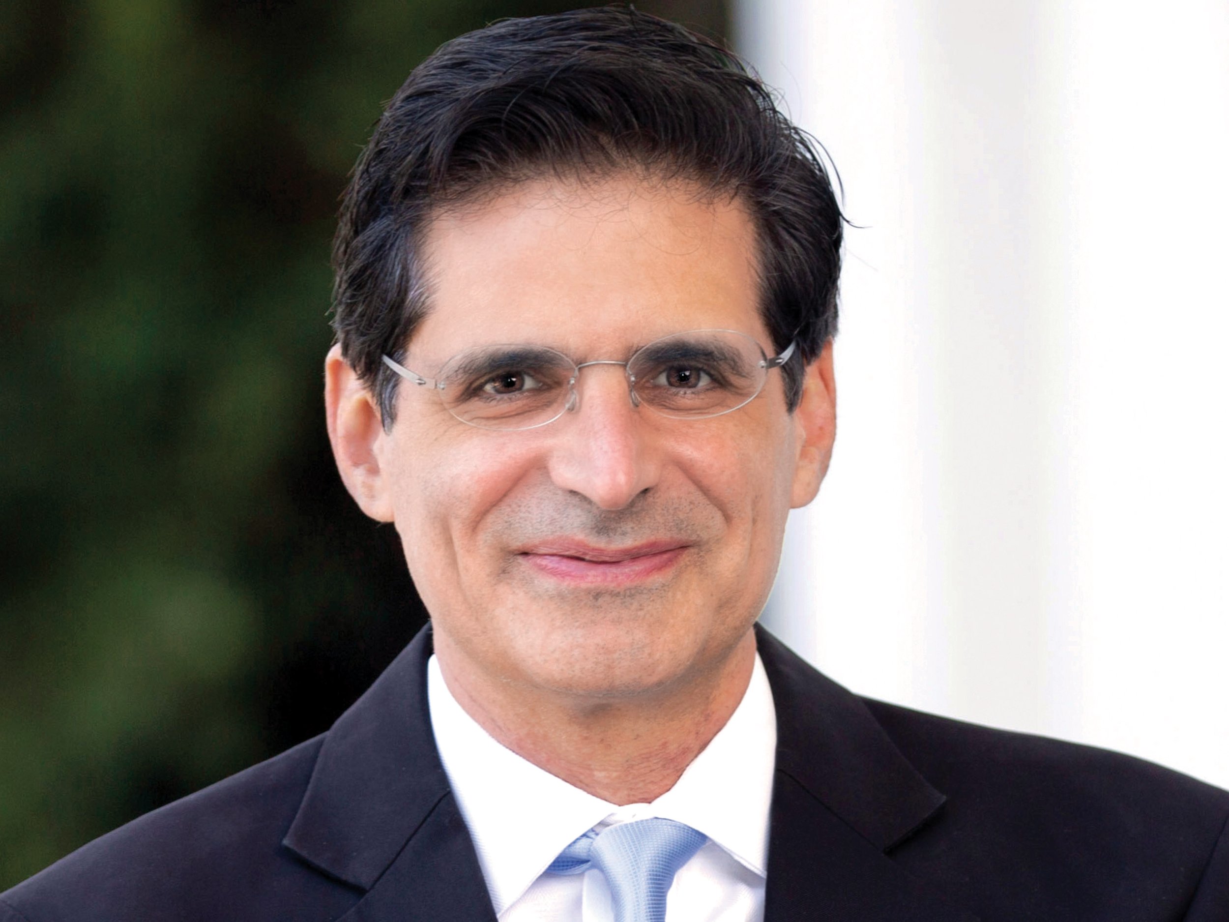 A man with glasses, dark hair, dressed in a black suit and white shirt, smiling outdoors with a blurred green background.