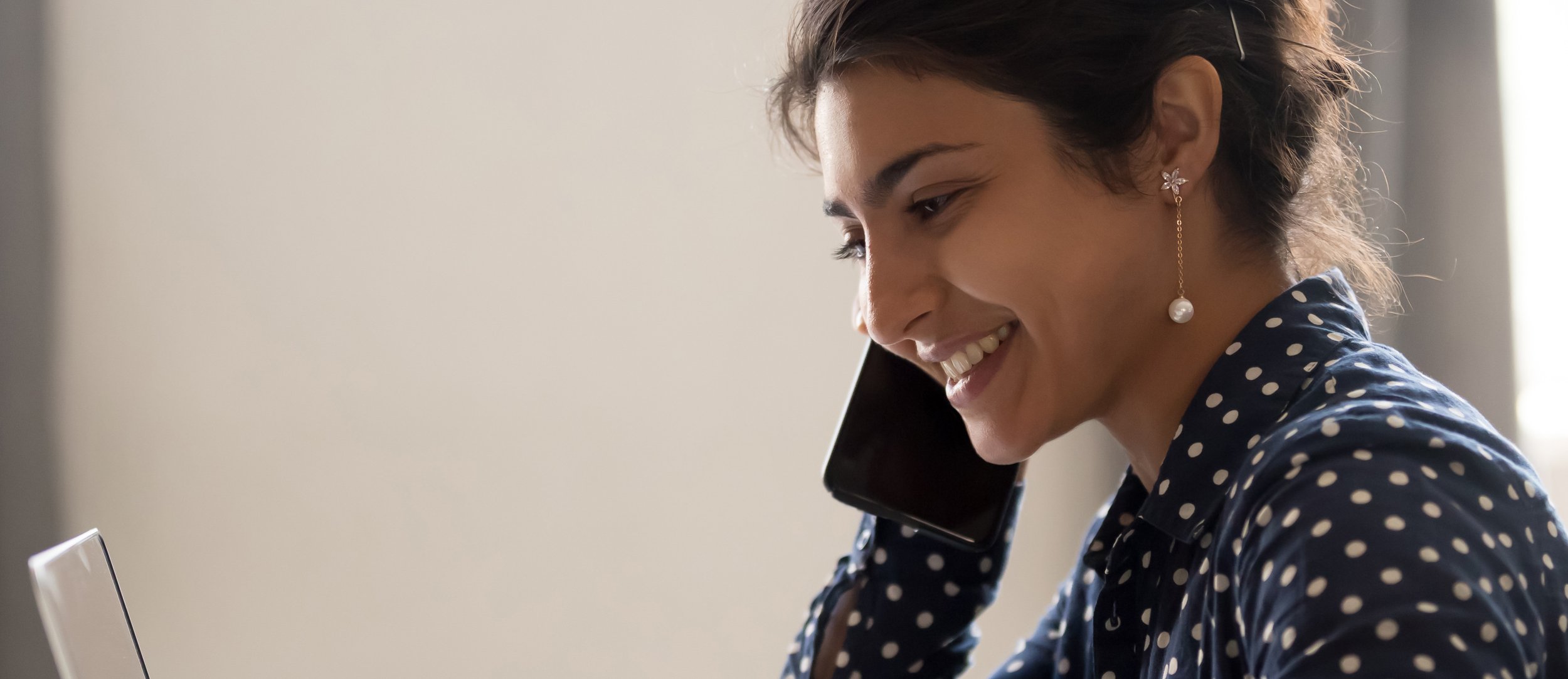 Smiling woman with dark hair and earrings talking on a black phone, wearing a blue polka dot shirt.