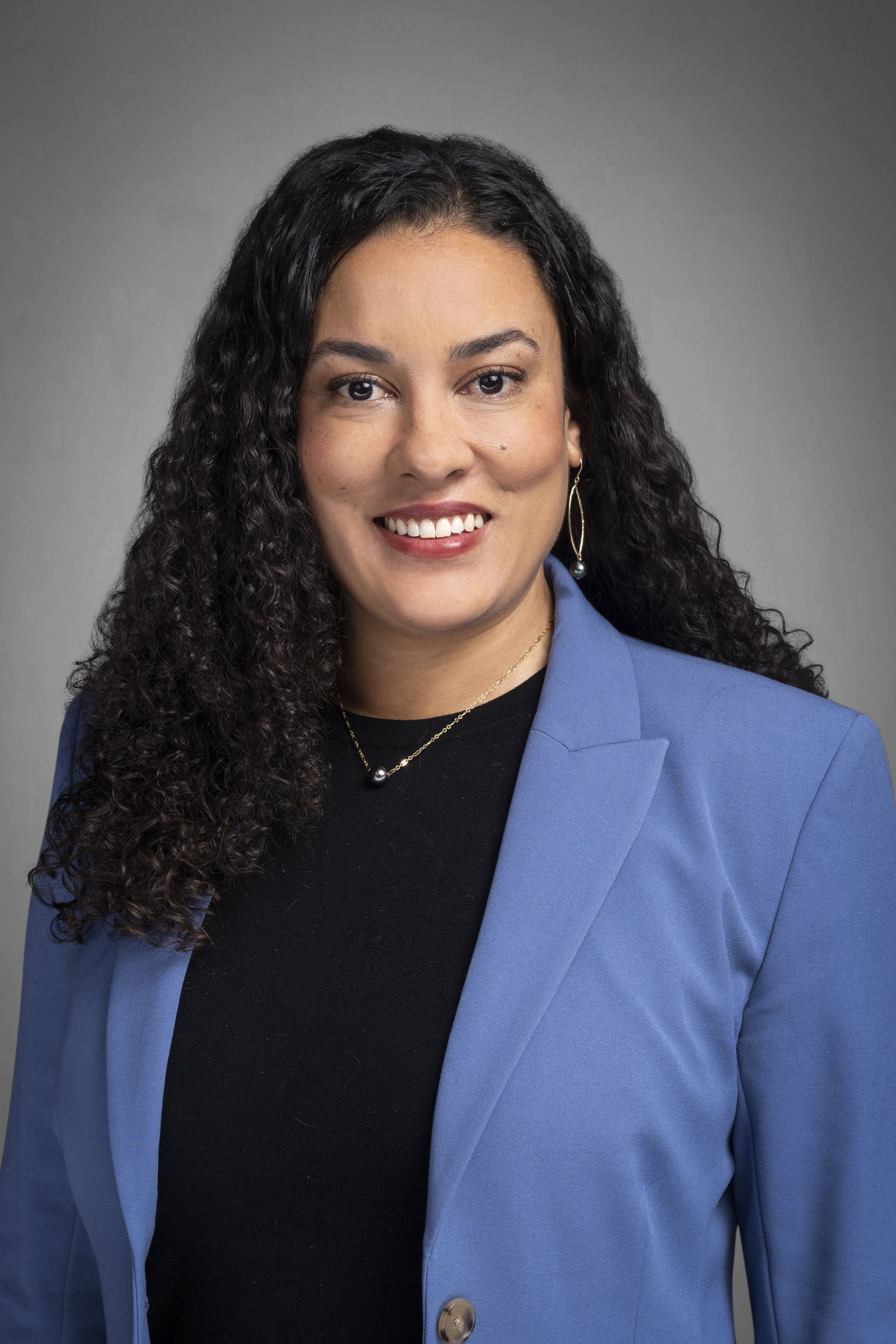 Professional headshot of a woman with curly dark hair, smiling, wearing a blue blazer, black top, and jewelry, against a gray studio background.
