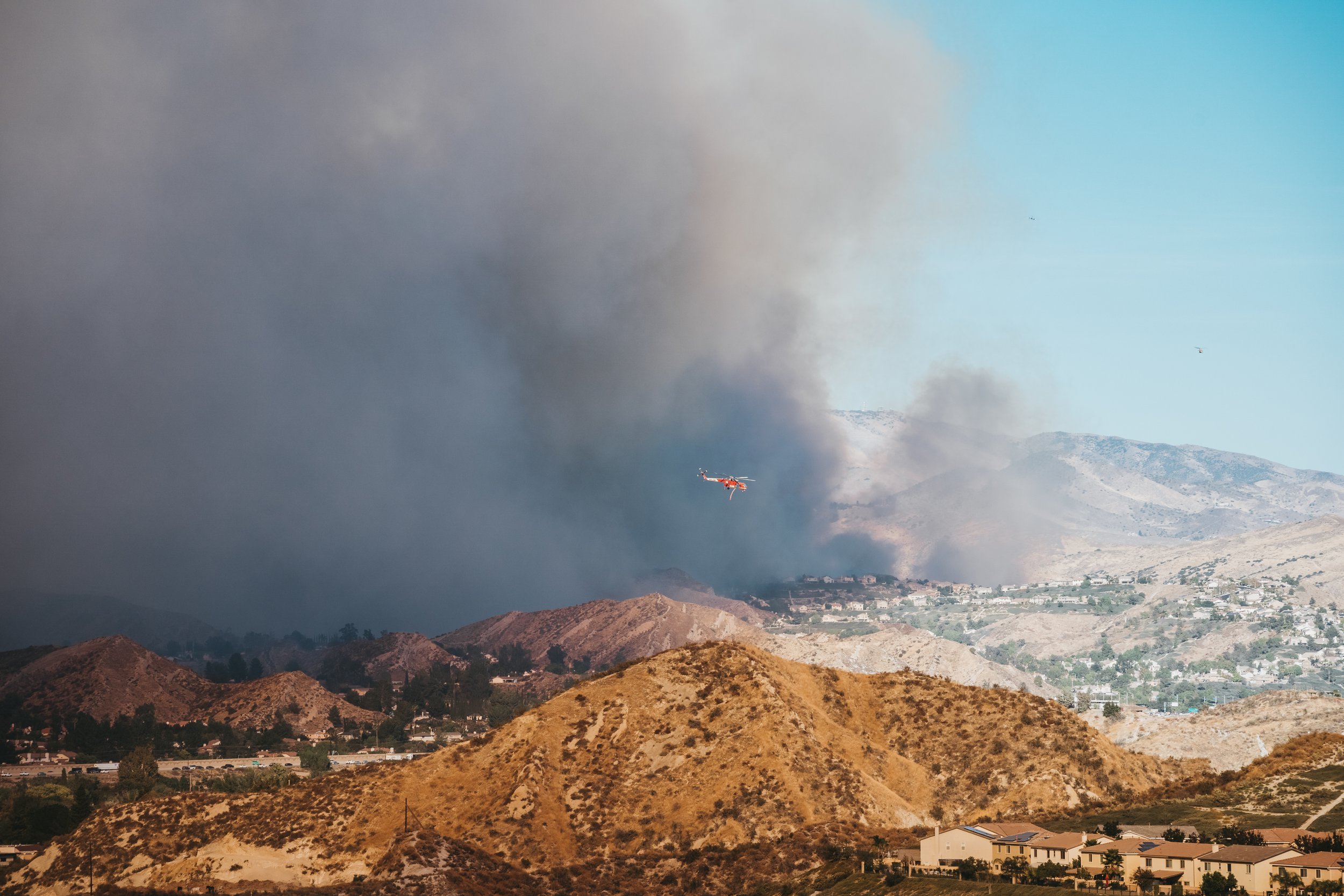 A helicopter flying over dry, brown hills with smoke and flames in the distance, suggesting a wildfire near a residential area.