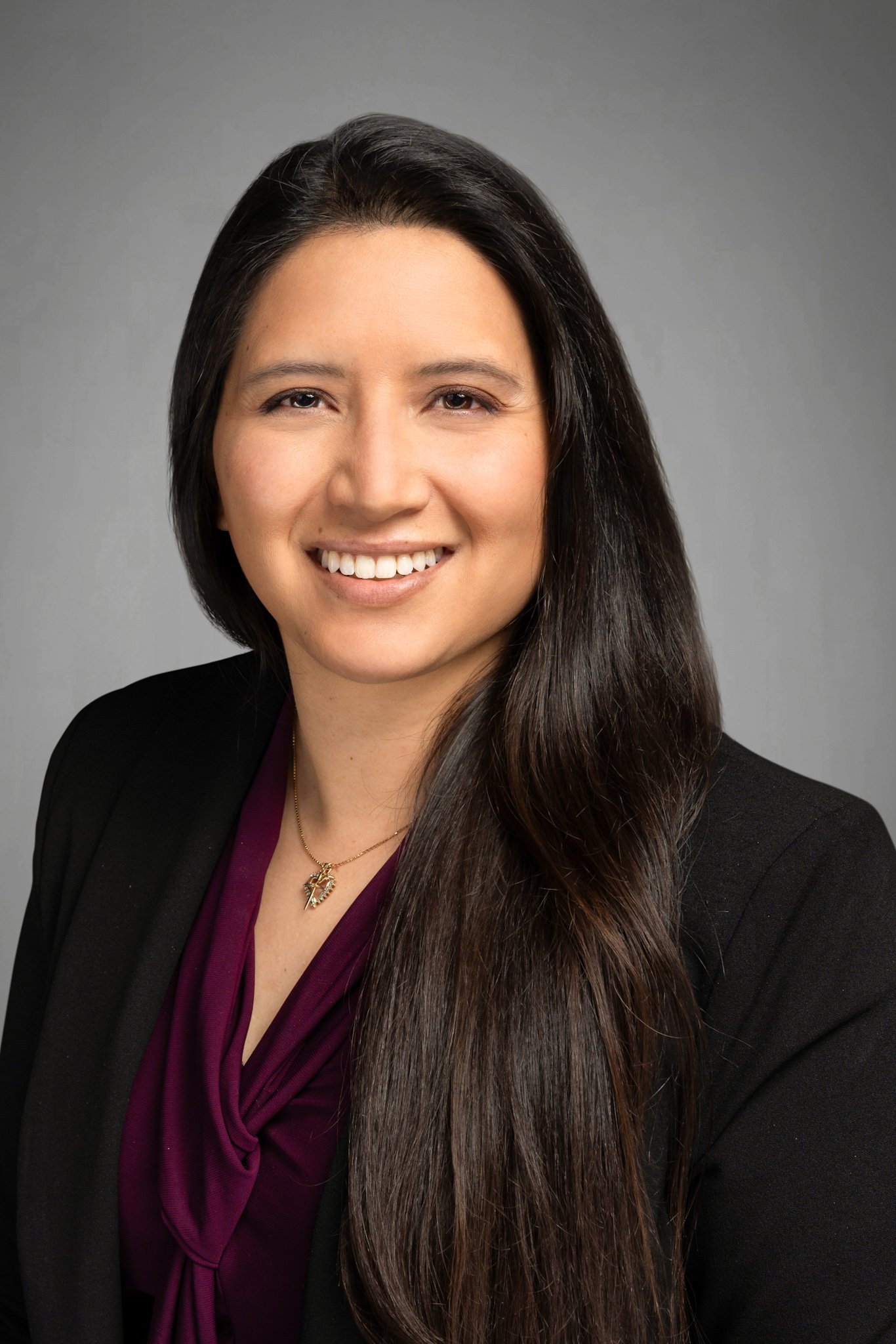 Professional headshot of a woman with long dark hair, wearing a black blazer and a purple blouse, smiling at the camera against a gray background.