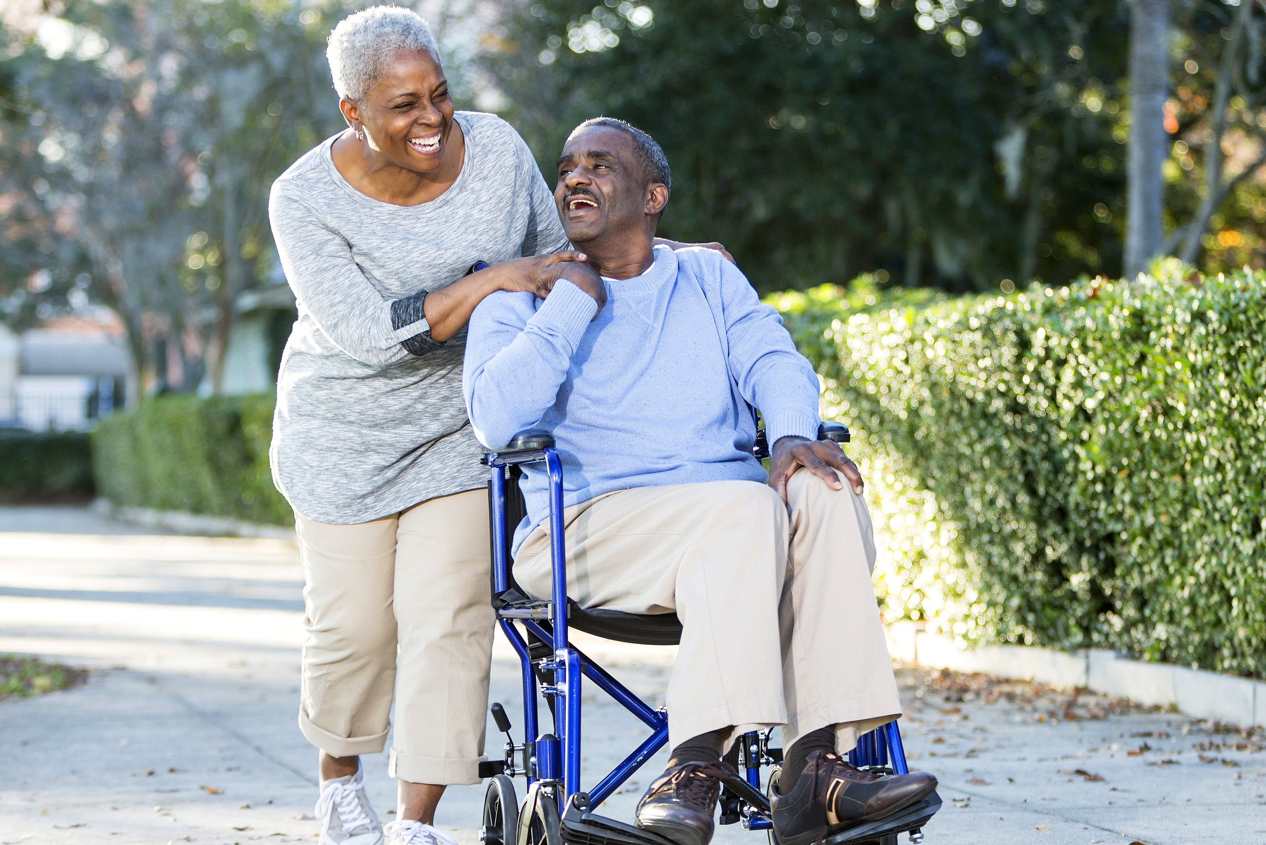 A smiling elderly woman helping a man in a wheelchair outdoors.