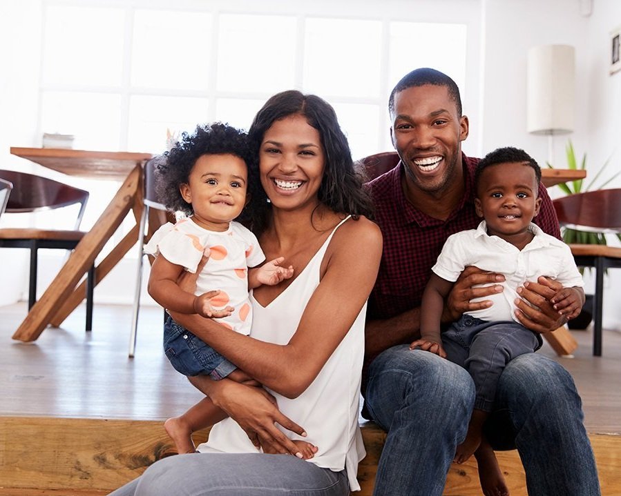 A happy family of four sitting on a wooden floor in a bright living room. The mother holds a smiling little girl with curly hair, while the father holds a cheerful young boy. All family members are smiling and looking at the camera.