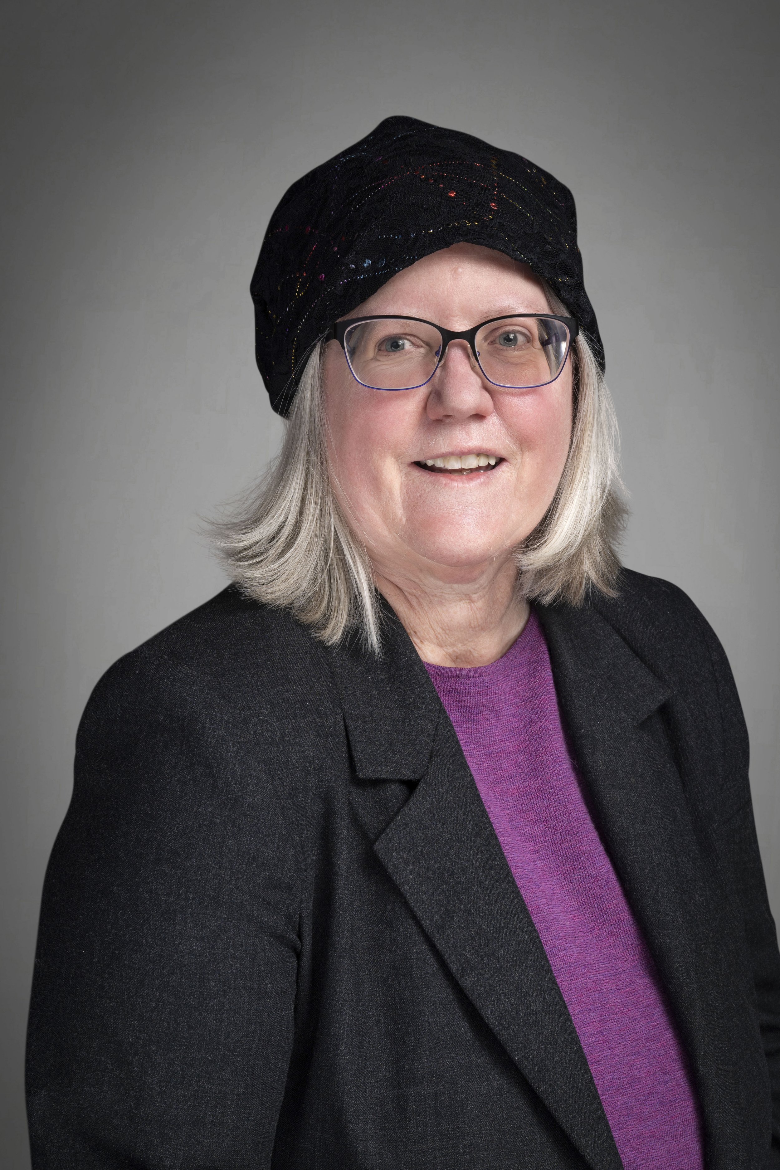 Portrait of a woman with shoulder-length gray hair, wearing glasses, a black blazer, a purple top, and a black headscarf with multicolored streaks, smiling at the camera.
