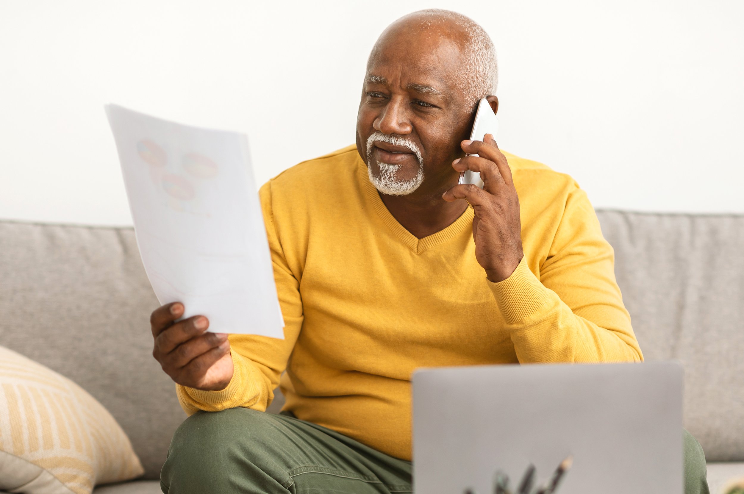 An older man with gray hair and beard wearing a yellow sweater, sitting on a couch and talking on the phone while holding a few sheets of paper.